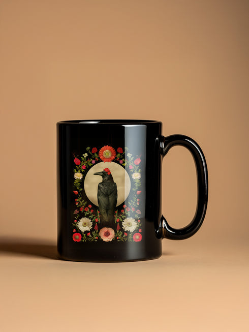 A black ceramic mug featuring a gothic design of a raven against a golden full moon, framed by a floral wreath of coral-colored daisies, white chrysanthemums, and delicate botanical elements. The design includes rich green foliage and small red buds arranged in a symmetrical pattern. The mug has a classic C-shaped handle with a glossy finish.